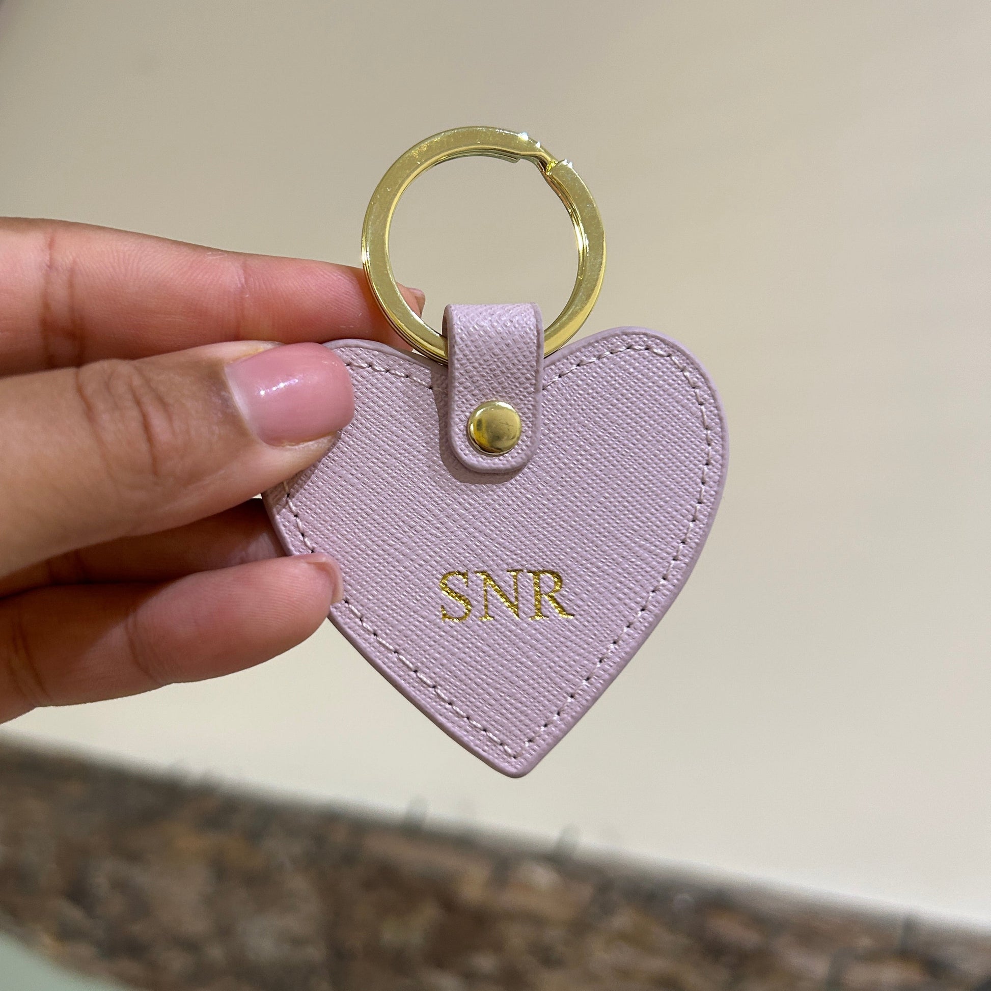 Hand holding a pink heart-shaped keychain with 'SNR' initials against a neutral background