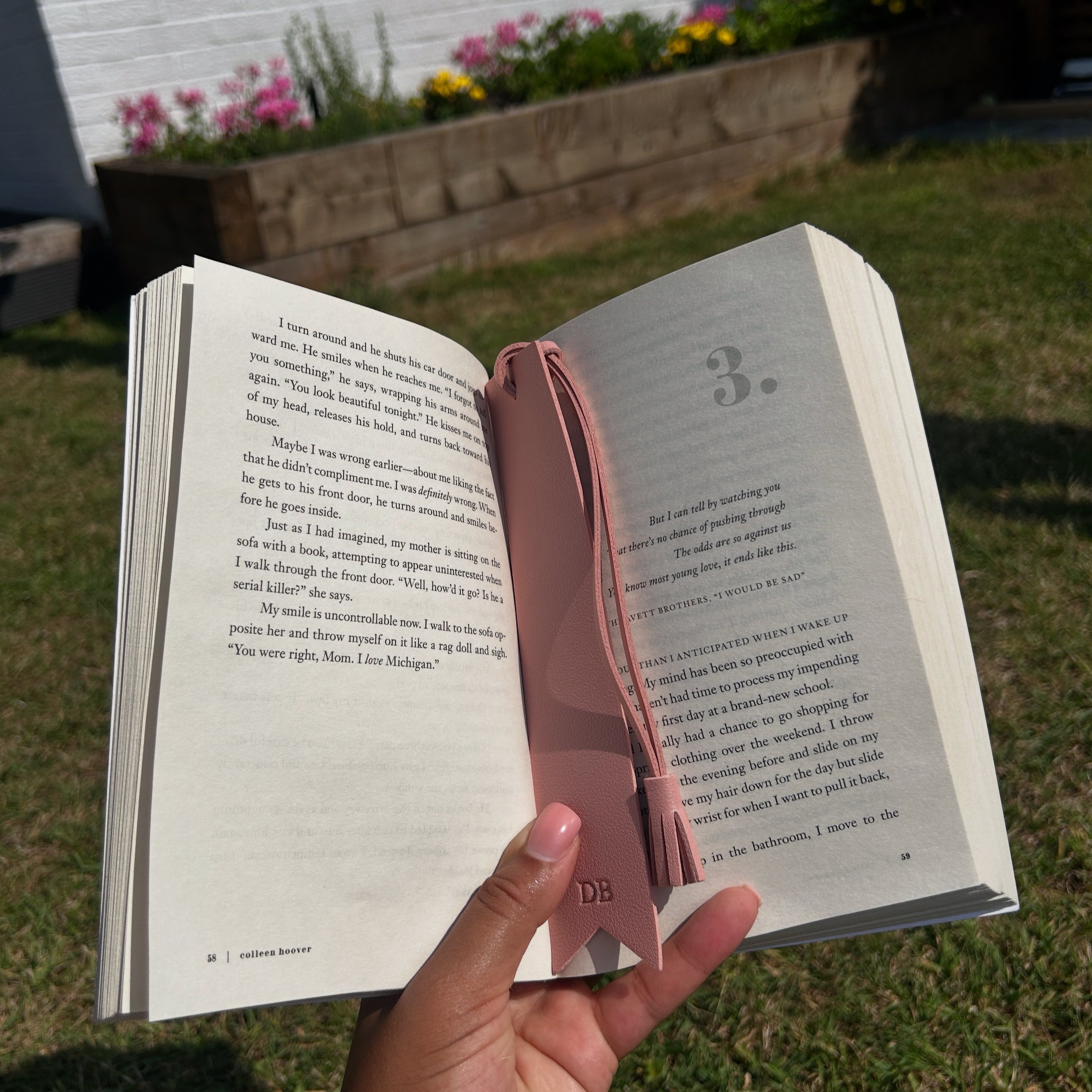 Hand holding an open book with a pink bookmark against a garden background