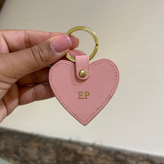Pink heart-shaped keychain with gold ring held by a hand against a neutral background