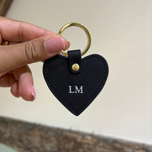 Black heart-shaped keychain with gold ring held by a hand against a neutral background