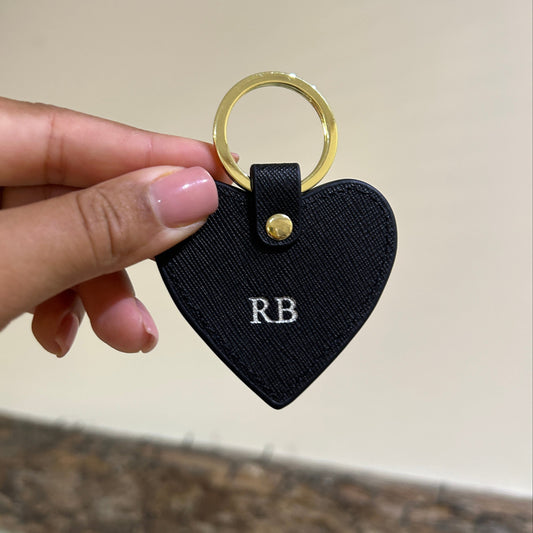 Black heart-shaped keychain with gold ring held by a hand against a neutral background