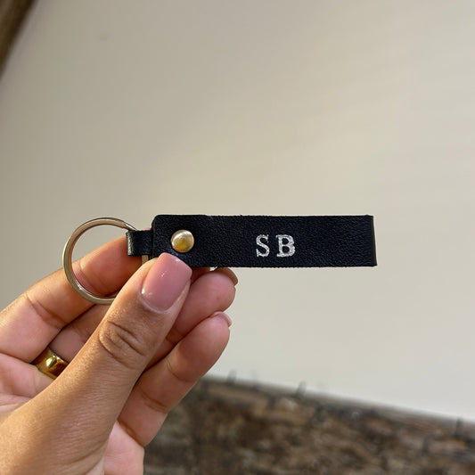 Hand holding a black leather key fob with initials 'SB' against a neutral background