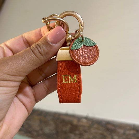Hand holding a brown leather keychain with a gold ring and green heart charm against a neutral background.