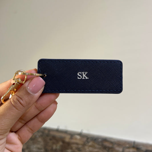 Navy keychain tag with 'SK' initials held by a hand against a neutral background