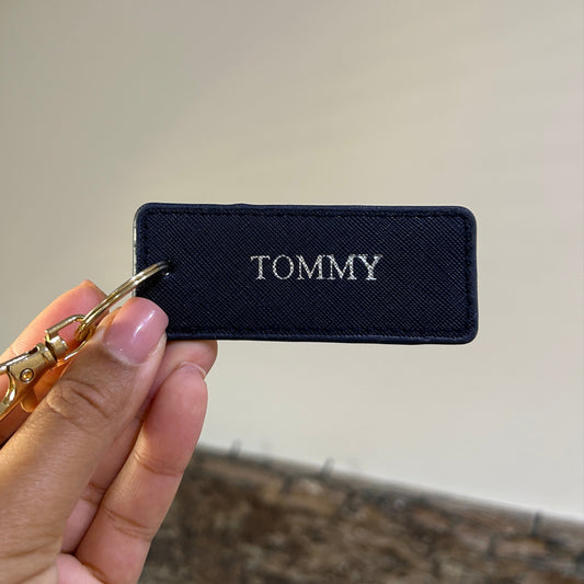 Hand holding a keychain with a Tommy written in silver against a neutral background