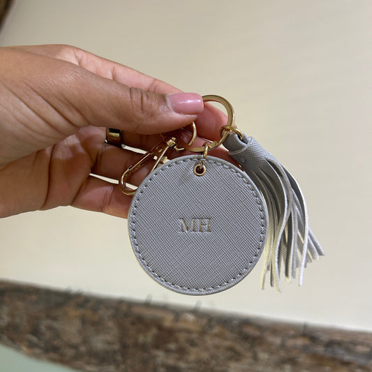 Round gray keychain with tassel held by a hand against a neutral background