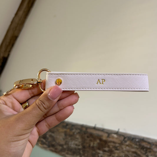 Hand holding a keychain with a strap and gold clasp against a neutral background