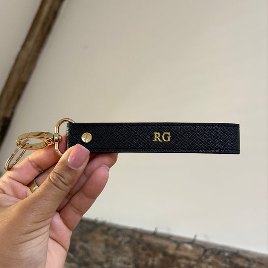 Black keychain strap with gold hardware held by a hand against a neutral background